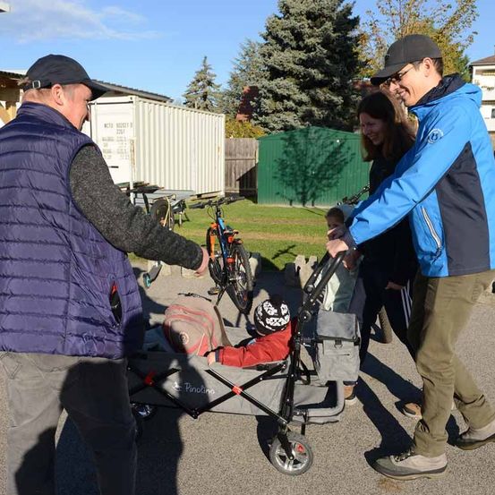 Zwei Männer, eine Frau und ein junges Kind stehen draußen in der Nähe von Fahrrädern. Der Mann in Blau schiebt einen Kinderwagen mit einem Kind darin.