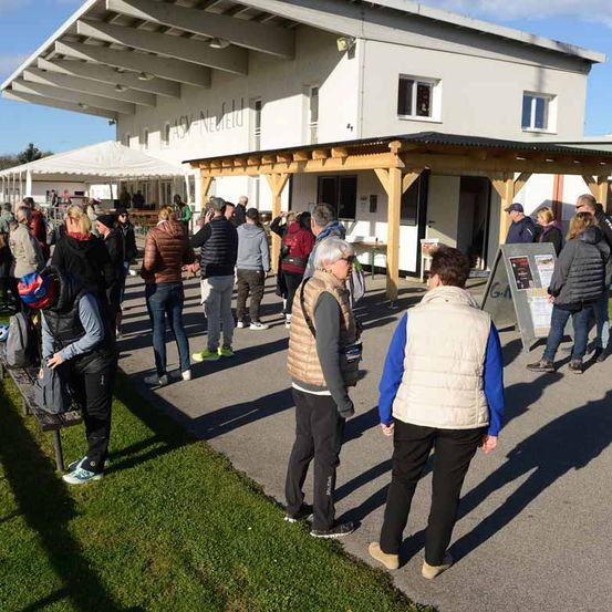 Eine Gruppe von Menschen steht in einer Schlange vor einem Gebäude mit dem Schild 'Easy-Nellei'. Das Gebäude hat weiße Wände und mehrere Fenster. Die Menschen tragen Winterkleidung und einige haben Taschen bei sich.