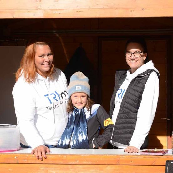 Drei Frauen stehen lächelnd in einer hölzernen Kabine. Ein junges Mädchen in einer blauen Mütze und einem Schal steht zwischen zwei Erwachsenen. Hinter ihnen befindet sich eine Holzwand mit einer Aufbewahrungsbox und einer hängenden Jacke.