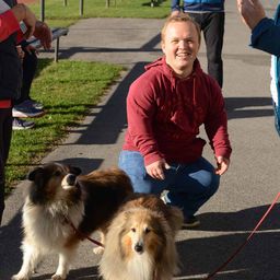 Ein Mann kniet sich hin und lächelt, während er zwei Collies an der Leine führt. Eine weitere Person steht hinter ihm.