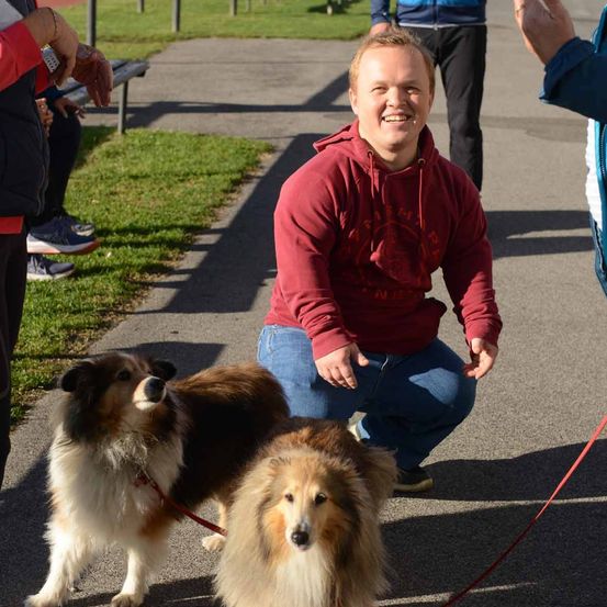 Ein Mann kniet sich hin und lächelt, während er zwei Collies an der Leine führt. Eine weitere Person steht hinter ihm.