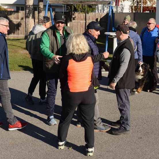 Mehrere Menschen stehen im Freien. Eine Frau mit orangem Weste spricht mit einem Mann in einer Mütze. Ein Hund läuft hinter ihnen.