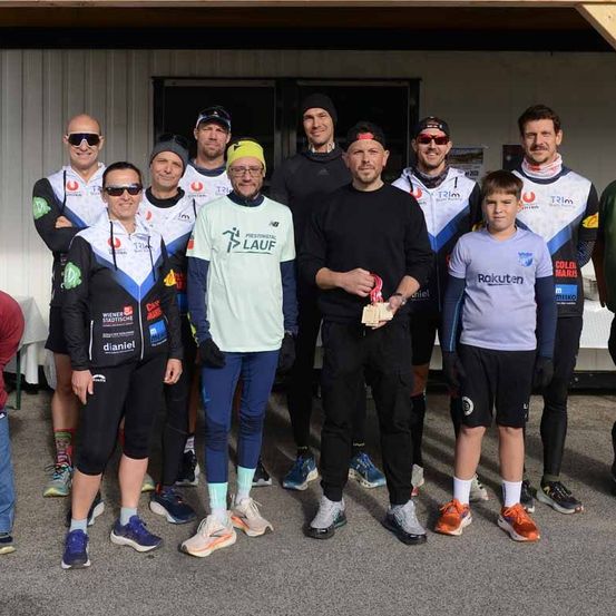 Eine Gruppe von Athleten, darunter zwei Frauen, posiert für ein Foto. Sie tragen Sporttrikots, Sonnenbrillen und Turnschuhe. Ein Mann in der Mitte hält eine Medaille. Hinter ihnen steht ein Junge mit einem weißen Tisch.
