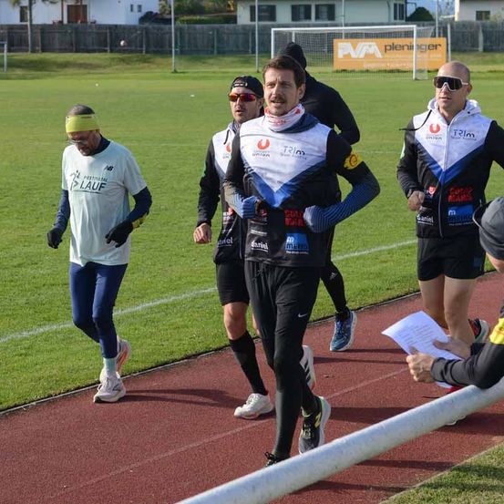 Mehrere Menschen joggen auf einer Laufbahn, darunter ein Mann mit einer gelben Kopfband. Ein Mann mit einem Papier steht rechts. Ein Fußballtor ist in der Ferne.