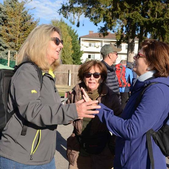 Drei Frauen in Jacken und mit Sonnenbrillen unterhalten sich im Freien. Eine trägt einen Rucksack, eine andere einen roten Rucksack. Hinter ihnen steht ein Holzzaun und Bäume, und ein Haus ist sichtbar.