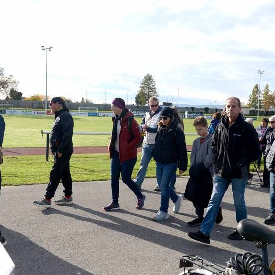 Eine Gruppe von Menschen geht auf einem Weg in einer Freiluftsportanlage. Sie sind in Winterkleidung gekleidet, einige tragen Tassen. Im Hintergrund ist ein Rasenfeld und ein Fußballfeld zu sehen.
