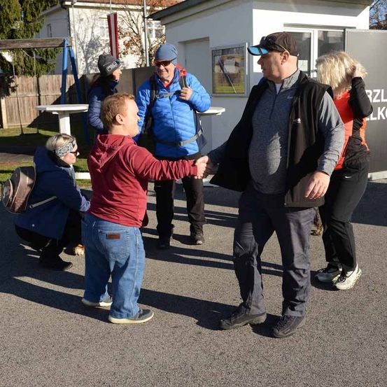 Eine Gruppe von Menschen steht draußen auf einem Parkplatz, zwei Männer schütteln sich die Hände, während andere hinter ihnen stehen, einer hält einen Rucksack.