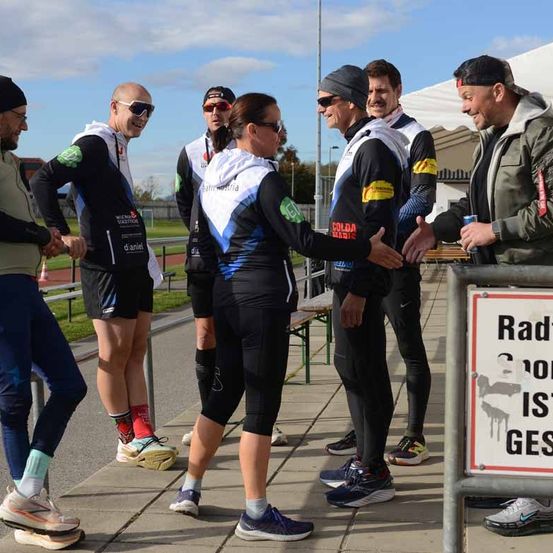Eine Gruppe von Athleten in Sportbekleidung schüttelt sich Hände an einer Laufbahn. Einer trägt ein weißes und blaues Shirt, der andere ein schwarzes und gelbes Shirt. Ein Schild steht 'Rad 15 IS GES'.