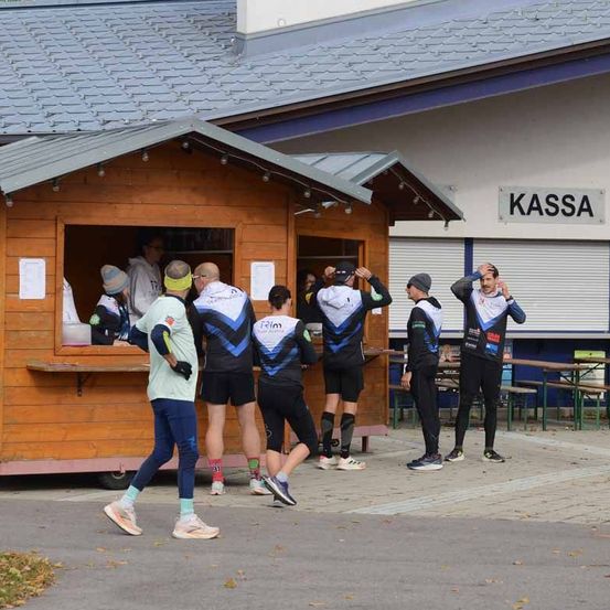 Eine Gruppe von Athleten in Radsportbekleidung steht vor einem Holzhäuschen, möglicherweise in Vorbereitung auf einen Wettkampf.