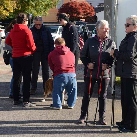 Eine Gruppe von Menschen, einige mit Brille, steht auf einem Parkplatz. Ein Mann beugt sich vor, während ein Hund zwischen ihnen hindurchgeht. Zwei Frauen halten Wanderstöcke und unterhalten sich miteinander.