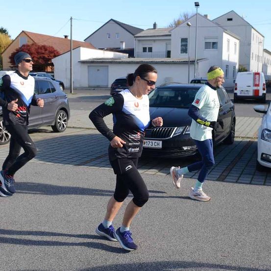 Drei Läufer joggen auf einem Parkplatz. Eine Frau führt, hinter ihr folgen ein Mann und eine weitere Frau. Dahinter sind mehrere Autos geparkt, und ein Lieferwagen ist sichtbar.