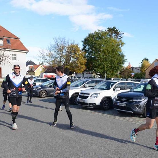 Teilnehmer laufen auf einer Straße und tragen Sportausrüstung. Dahinter sind mehrere Autos geparkt, darunter ein weißer Chevrolet und ein grauer Volkswagen. Gebäude und Bäume sind im Hintergrund.