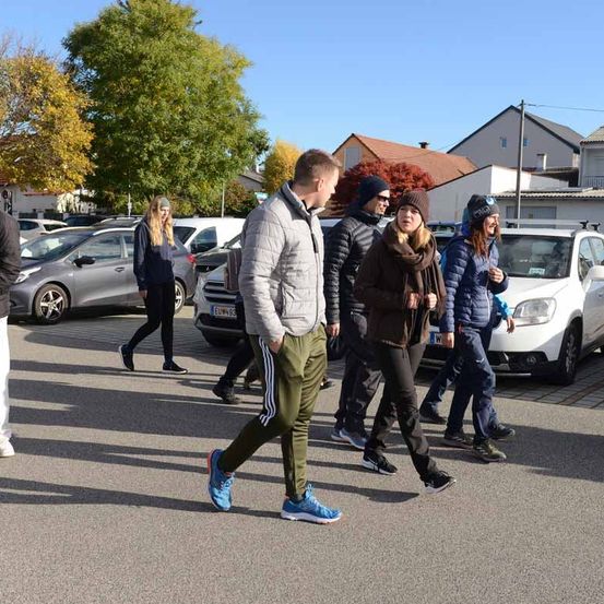 Eine Gruppe von Menschen läuft auf der Straße in einem Parkplatz. Sie tragen alle Winterkleidung. Es sind Autos am Straßenrand geparkt.