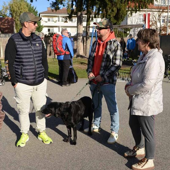 Drei Personen und ein schwarzer Hund unterhalten sich auf einem Parkplatz. In der Nähe stehen einige Personen, und Fahrräder sind geparkt. Im Hintergrund befindet sich ein Spielplatz mit Schaukeln.