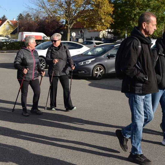 Zwei Frauen und ein Mann gehen mit ihren Wanderstöcken auf einem Parkplatz. Die Frauen tragen eine Brille und eine Jacke. Hinter ihnen sind Autos auf einem Parkplatz geparkt.