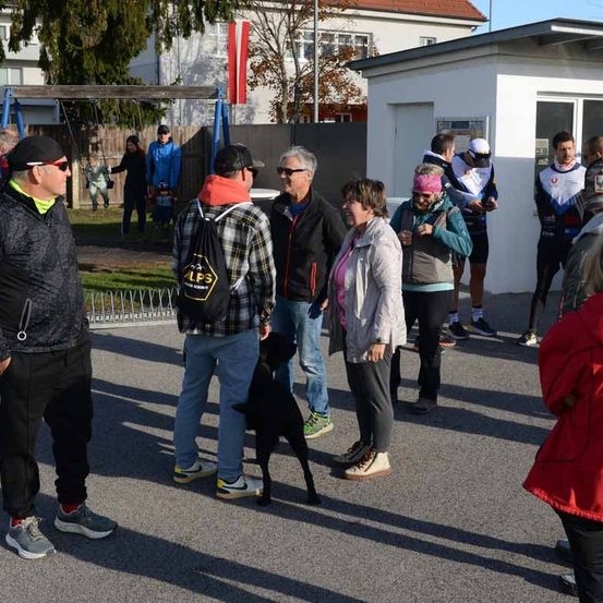 Eine Gruppe von Menschen in Outdoor-Kleidung versammelt sich auf einem Parkplatz, im Hintergrund ein Spielplatz. Eine Person trägt einen Rucksack und eine andere hat einen Hund.