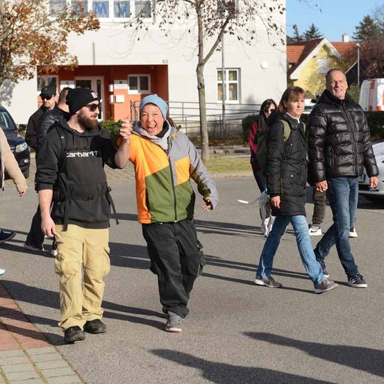 Mehrere Menschen gehen auf einer Straße, gesäumt von Autos, Gebäuden und Bäumen. Eine Frau lacht und hält die Hand eines Mannes.