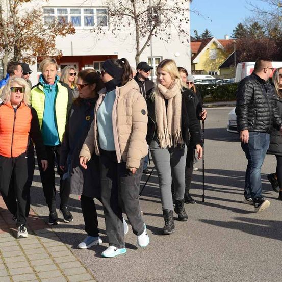 Eine Gruppe von Menschen in Winterkleidung geht auf einer Straße. Einige tragen Sonnenbrillen und haben Stöcke dabei. Sie stehen vor einem Gebäude mit Glasfenstern.