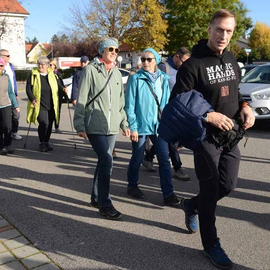 Eine Gruppe von Menschen geht auf einem asphaltierten Weg. Sie sind in verschiedene Jacken gekleidet, einige mit Schals, und einige tragen Stöcke. Ein Mann hält eine blaue Jacke. Bäume und Gebäude sind im Hintergrund.