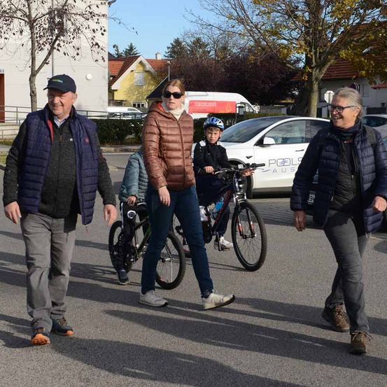Vier Personen gehen mit Fahrrädern auf einem Parkplatz. Ein weißes Auto ist hinter ihnen geparkt. Zwei Kinder fahren Fahrräder. Bäume und Häuser sind im Hintergrund.