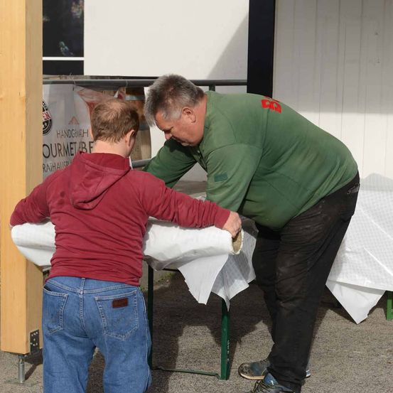 Zwei Männer in grünen und roten Jacken arbeiten zusammen, um einen Tisch mit weißer Tischdecke im Freien zu decken.