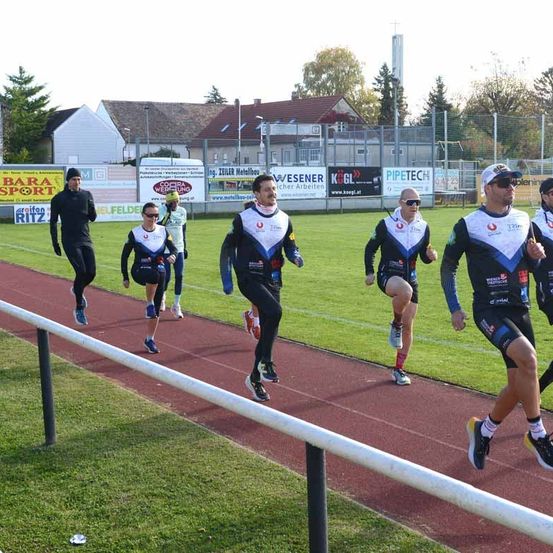 Eine Gruppe von Läufern, in identischen Outfits gekleidet, sprintet auf einer Laufbahn. Dahinter erstreckt sich ein Grasfeld, mit einem Zaun und Gebäuden in der Ferne.