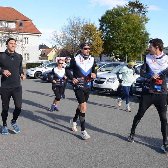 Eine Gruppe von Läufern joggt im Freien. Sie tragen Trikots und Turnschuhe. Es sind geparkte Autos und Bäume im Bereich. Dahinter befindet sich ein Gebäude mit einem roten Dach.