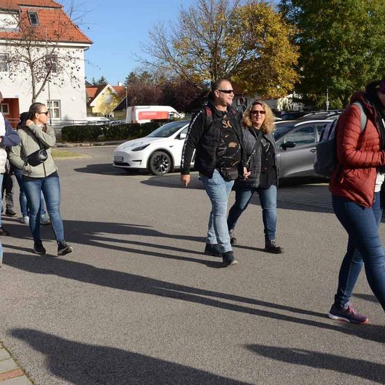Mehrere Menschen gehen auf einer Straße, hinter ihnen ist ein weißer Tesla Model 3 geparkt. Sie tragen Freizeitkleidung, und einige tragen Rucksäcke. Das Gebiet ist von Bäumen umgeben, und im Hintergrund sind Gebäude zu sehen.