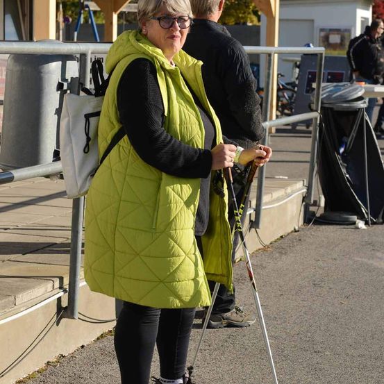 Eine Frau in einer leuchtend gelben Jacke steht auf einem Weg mit Trekkingstöcken, ein Mann hinter ihr. In der Nähe ist ein Mülleimer mit Säcken gefüllt.