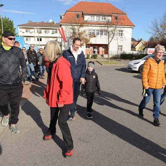 Eine Gruppe von Menschen geht auf einer Straße. Einige tragen Jacken und Turnschuhe. Ein Junge führt den Weg. Hinter ihnen sind Gebäude, Autos und Bäume zu sehen.