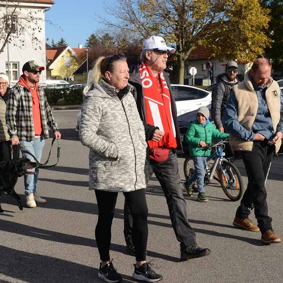 Eine Gruppe von Menschen geht auf einer Straße, einige mit Haustieren. Ein Mann mit Schal und eine Frau mit Pferdeschwanz stehen in der Mitte, ein junger Junge fährt in der Nähe ein Fahrrad. Bäume und Häuser sind im Hintergrund.