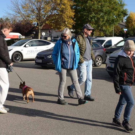 Eine Familie mit einem kleinen Hund geht auf einem Parkplatz, wo mehrere Autos geparkt sind. Die Frau trägt eine Sonnenbrille und eine blaue Jacke. Der Mann neben ihr trägt eine grüne Jacke. Der Junge neben ihnen trägt eine Mütze.