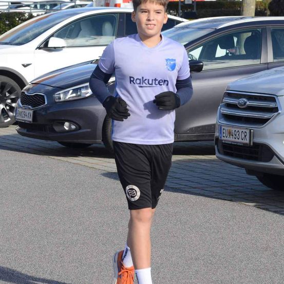 Ein junger Junge in einem Rakuten-Trikot und schwarzen Shorts läuft auf einer Straße, mit geparkten Autos im Hintergrund.