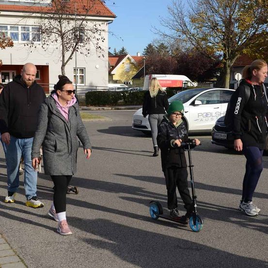 Eine Gruppe von Menschen, darunter ein Kind auf einem Roller, geht eine Straße entlang. Hinter ihnen ist ein weißer Wagen geparkt. Gebäude mit roten Dächern befinden sich links, und ein Baum mit grünen Blättern rechts.