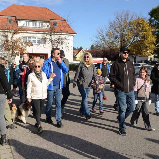 Eine Gruppe von Menschen, darunter Erwachsene und Kinder, geht auf einem Parkplatz. Einige tragen eine Brille und Turnschuhe. Ein Hund begleitet sie. Im Hintergrund steht ein Gebäude mit rotem Dach und Bäumen.