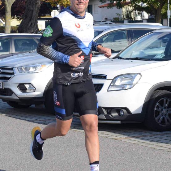 Ein Mann in Sportbekleidung läuft auf der Straße, mit geparkten Autos auf beiden Seiten. Er lächelt und gibt einen Daumen hoch.