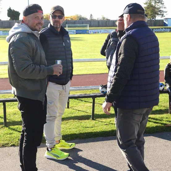 Drei Männer stehen auf einem Betonweg, möglicherweise bei einer Sportveranstaltung. Sie lächeln und schauen sich gegenseitig an. Einer von ihnen hält eine Dose. Hinter ihnen befindet sich ein grasbewachsenes Feld mit einem weißen Zaun und einem Fußballtor.