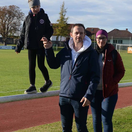 Ein Mann und eine Frau stehen auf einem Sportplatz mit einem Kind. Das Kind steht auf einem Bordstein, und der Mann hält eine Münze. Sie sind warm gekleidet in Jacken und Turnschuhen. Hinter ihnen befindet sich ein Rasenfeld mit einem Fußballtor.