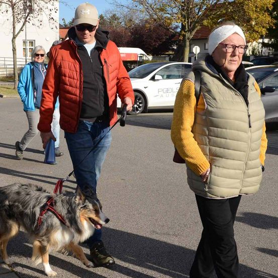 Zwei Personen gehen mit einem Hund auf einem Parkplatz, einer hält die Leine. Der Mann trägt eine rote Jacke und die Frau eine beige Weste. Sie werden von einer Frau in einer blauen Jacke begleitet. Im Hintergrund sind Autos geparkt.