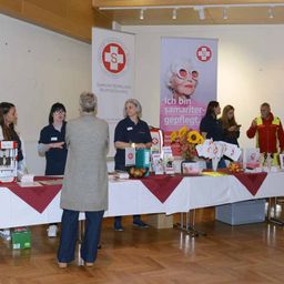Eine Gruppe von Menschen steht an einem Tisch mit verschiedenen Gegenständen, vielleicht bei einer Wohltätigkeitsveranstaltung. Hinter ihnen ist ein Banner mit einer Frau in Sonnenbrille angebracht.
