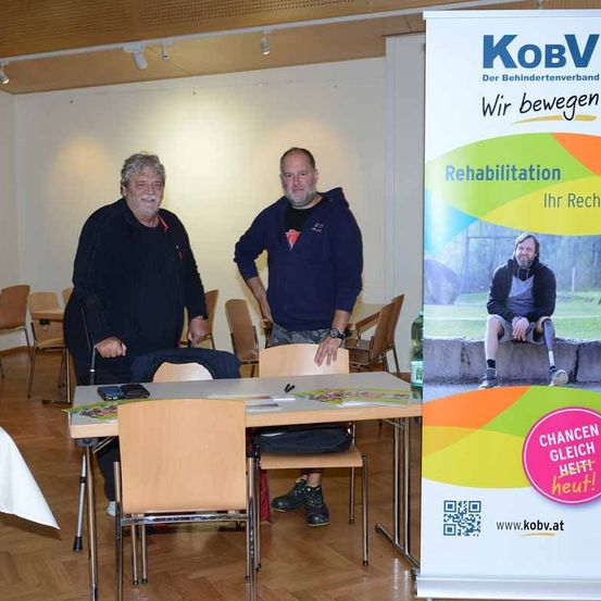 Zwei Männer stehen neben einem Tisch mit dem Schild 'Rehabilitation'. Sie befinden sich in einem Raum mit einem Banner und Stühlen.