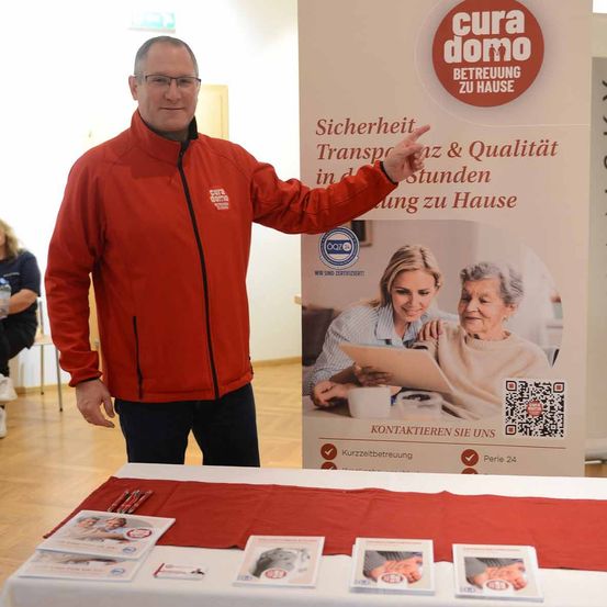 Ein Mann in einem roten Jacken steht neben einem Banner mit einer älteren Frau und einer jungen Frau. Unter dem Banner befindet sich ein Tisch mit Broschüren.