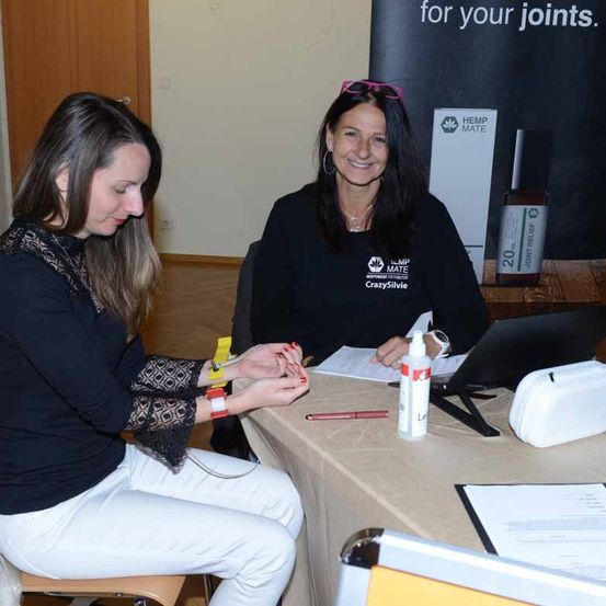 Zwei Frauen in einem Raum mit einem Banner, das 'für Ihre Gelenke' sagt. Eine Frau hält einen Stift, während die andere ein kleines Objekt hält.