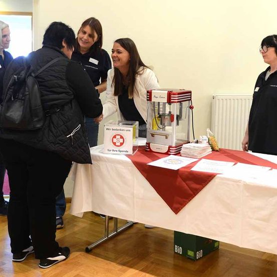 Mehrere Personen stehen um einen Tisch mit rot-weißem Tischtuch. Eine von ihnen lächelt und schaut eine andere Person an. Auf dem Tisch steht eine Box mit dem Text 'Wir bedanken uns für Ihre Spende' und eine Popcornmaschine.