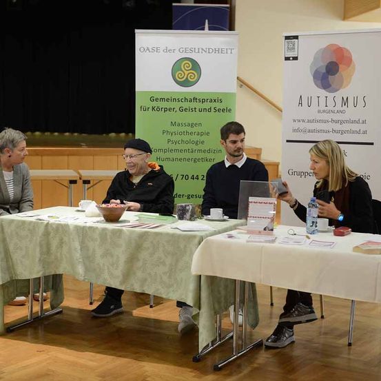 Vier Personen sitzen an einem Tisch in einem Raum. Hinter ihnen befinden sich Banner mit Texten über Autismus und Gemeinschaftspraxis. Die Leute scheinen in einer Diskussion vertieft zu sein.