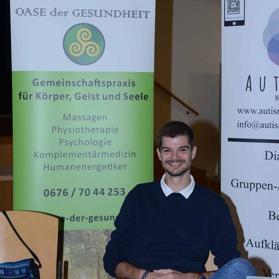 Ein Mann lächelt und sitzt vor einem Banner, das Dienstleistungen für Körper, Geist und Seele bewirbt. Das Banner hat ein Logo und beinhaltet Massage, Physiotherapie, Psychologie und Komplementärmedizin. Kontaktinformationen sind ebenfalls vorhanden.