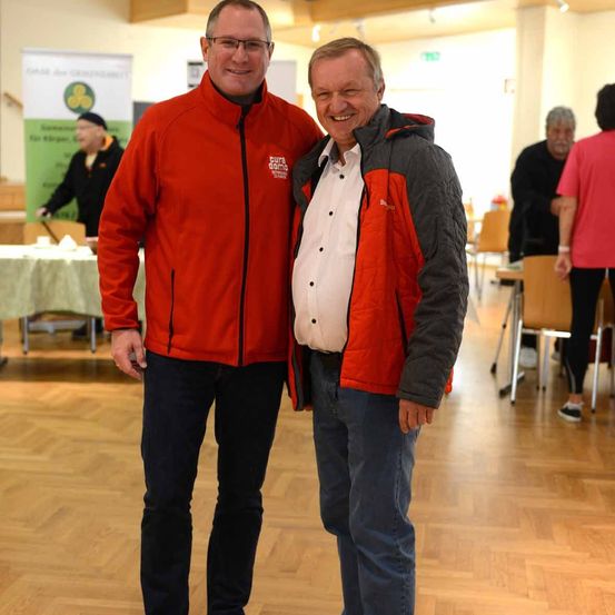 Zwei Männer in roten Jacken stehen lächelnd in einem Raum. Hinter ihnen geht eine Person in Schwarz. Es gibt Tische, Stühle und ein Banner im Raum.