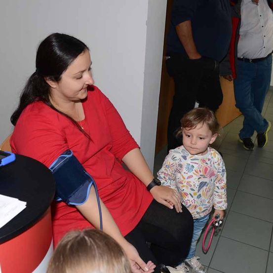 Eine Frau überprüft den Blutdruck eines kleinen Kindes mit einem Manschette, während ein Mann hinter ihnen steht.