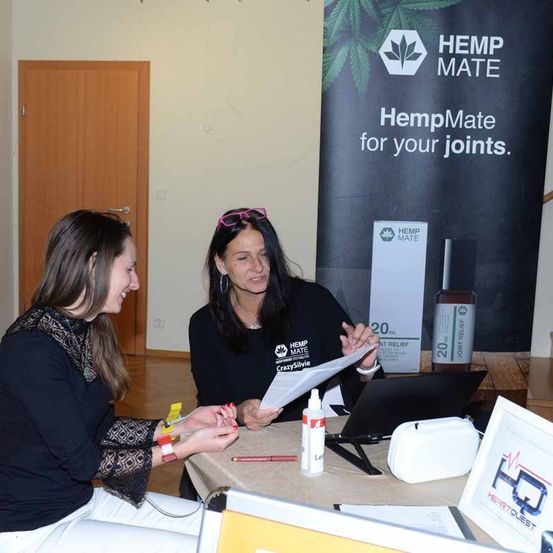 Zwei Frauen sitzen an einem Tisch mit Laptop, Papieren und einer Flasche. Sie tragen schwarze T-Shirts mit HempMate-Marke. Dahinter befindet sich ein HempMate-Banner und eine Holztür.