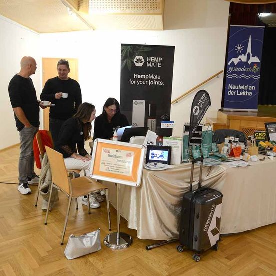 Eine Gruppe von Menschen versammelt sich um einen Tisch mit verschiedenen Gegenständen, wahrscheinlich bei einer HempMate-Veranstaltung. Zwei Frauen sitzen am Tisch, und ein Mann steht in der Nähe. Ein Banner für HempMate befindet sich hinter ihnen.